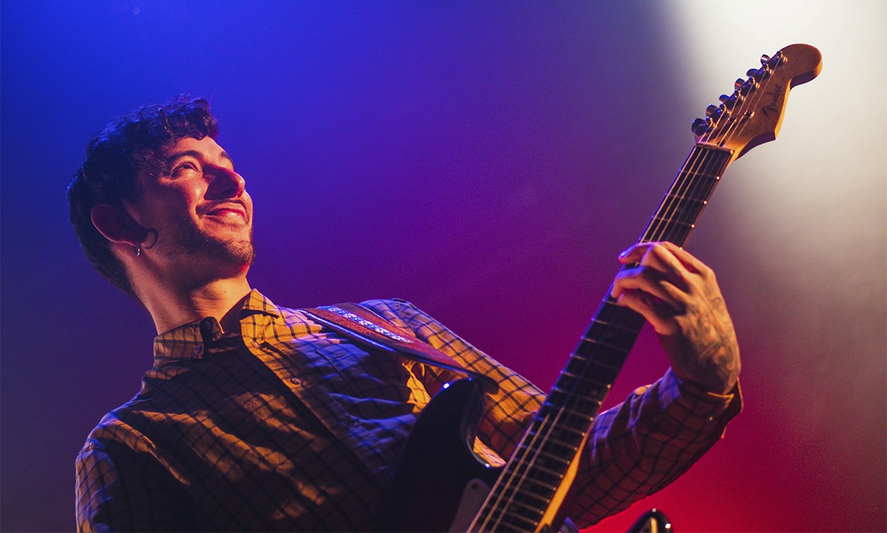 Guitarrista actuando en un concierto bajo luces azules, violetas y rojas.