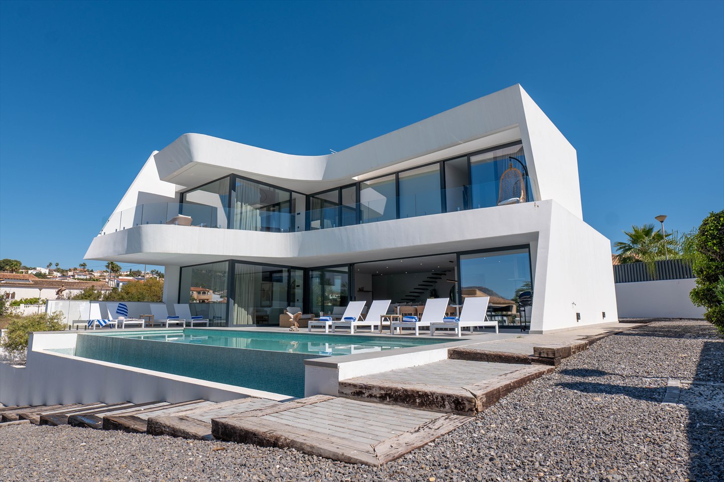 Villa contemporánea blanca con grandes ventanales y piscina exterior bajo un cielo azul.