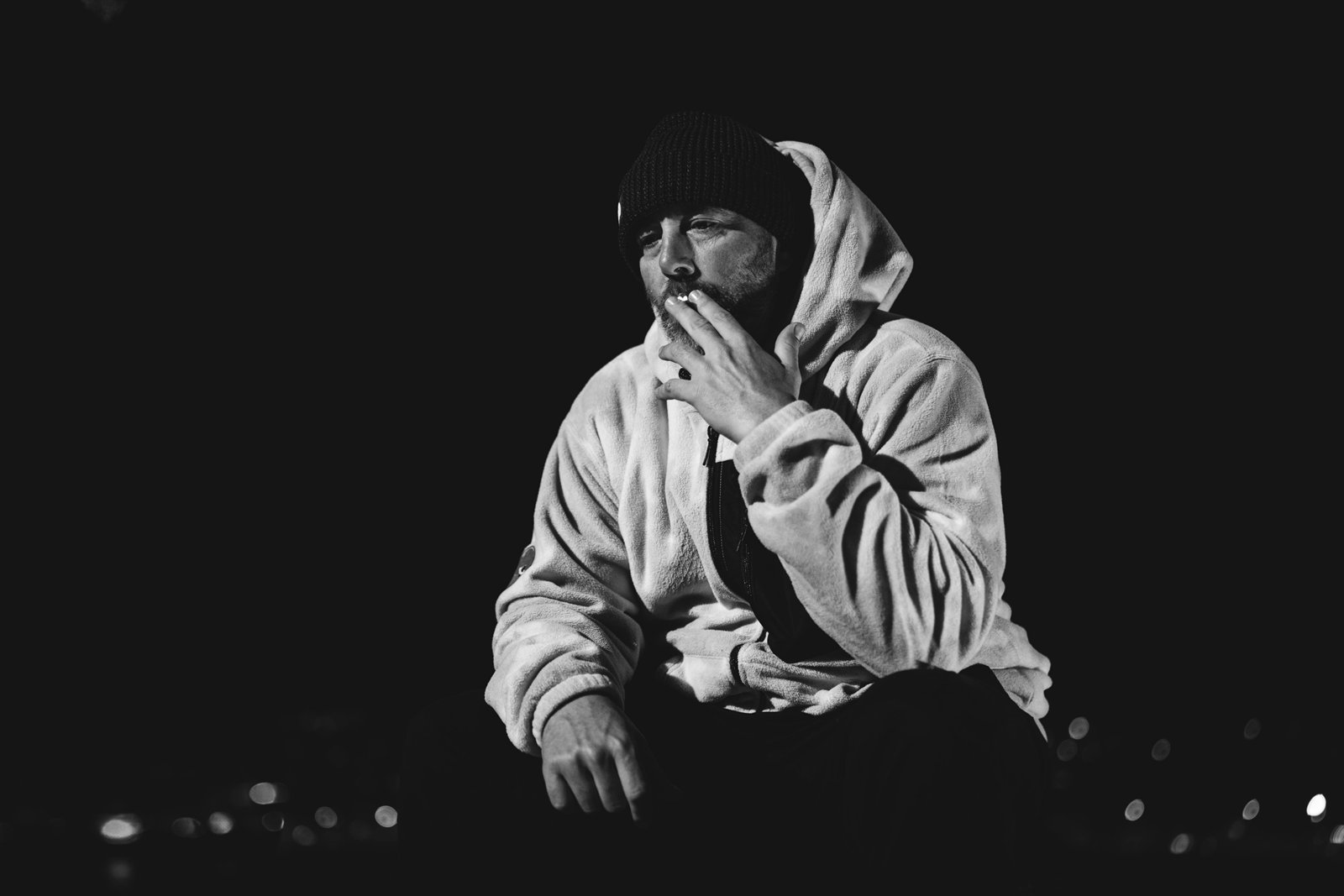 Retrato en blanco y negro de un hombre con gorro y sudadera sentado en la oscuridad.