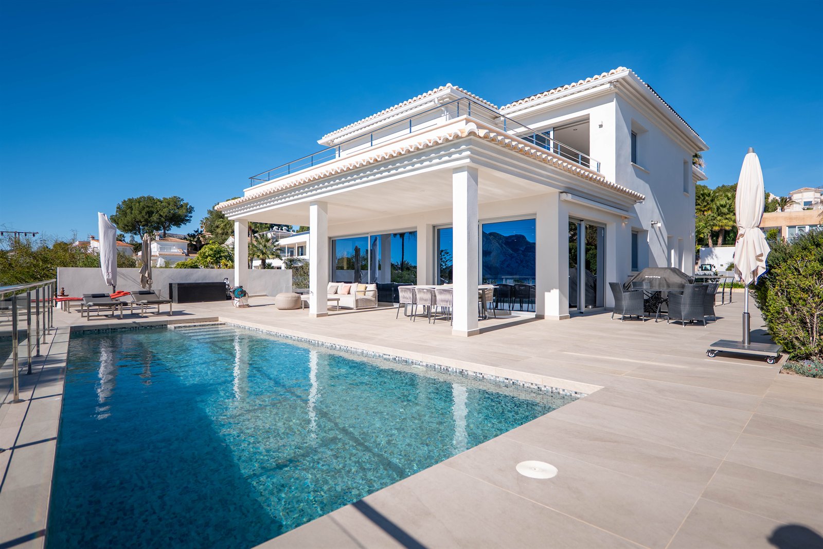 Villa blanca con terraza amueblada y piscina exterior bajo un cielo azul.