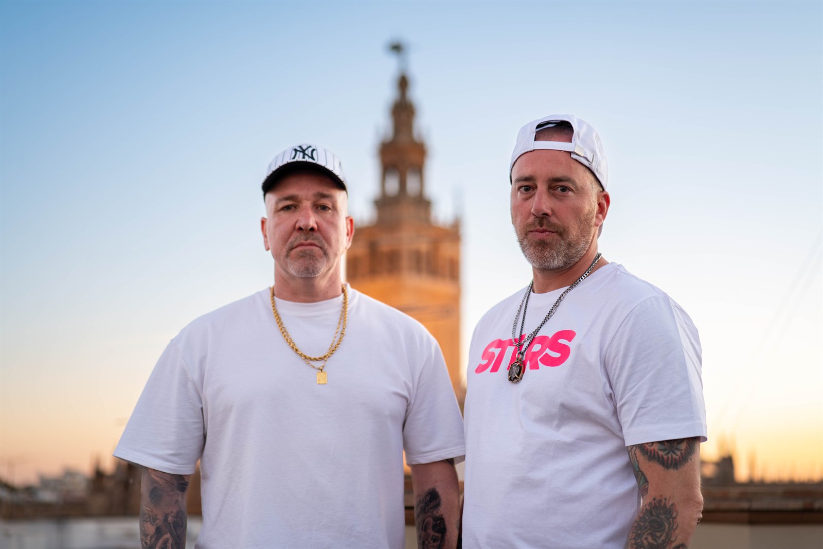 Dos hombres con camisetas blancas y gorras posan ante una torre al atardecer.