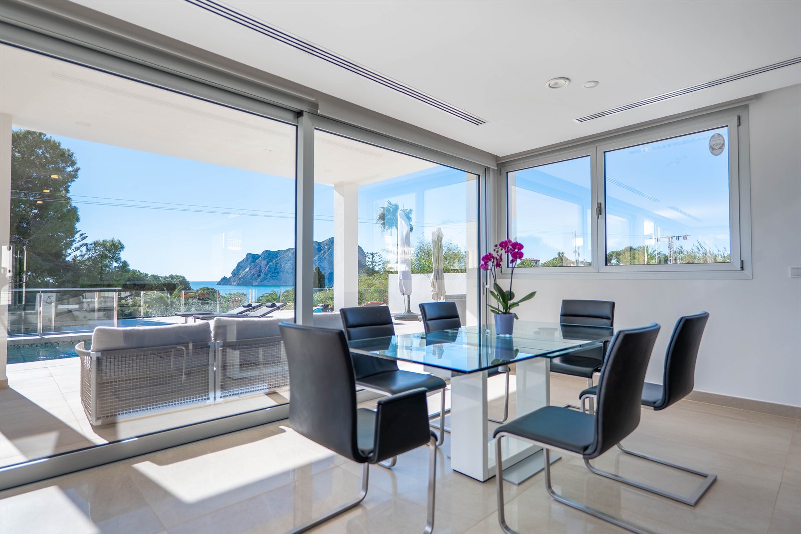 Comedor luminoso con grandes ventanales abiertos hacia una terraza, piscina y montaña.