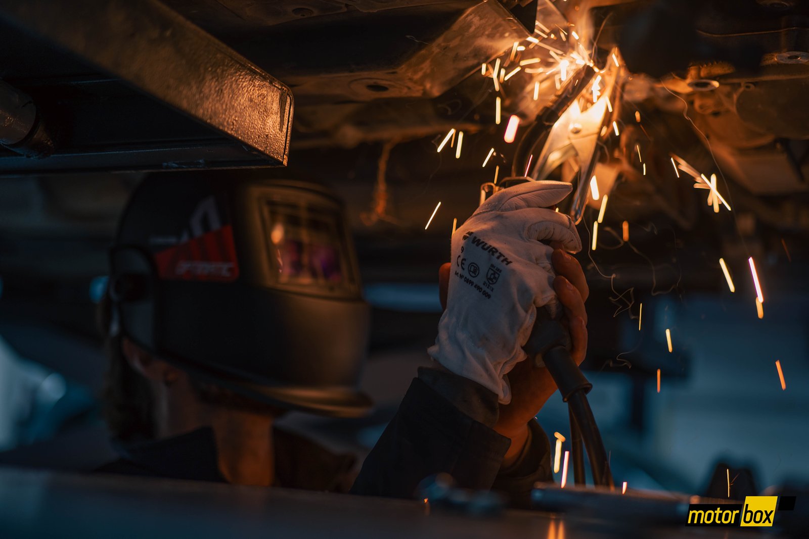 Mecánico con careta y guantes trabajando bajo un vehículo entre chispas.