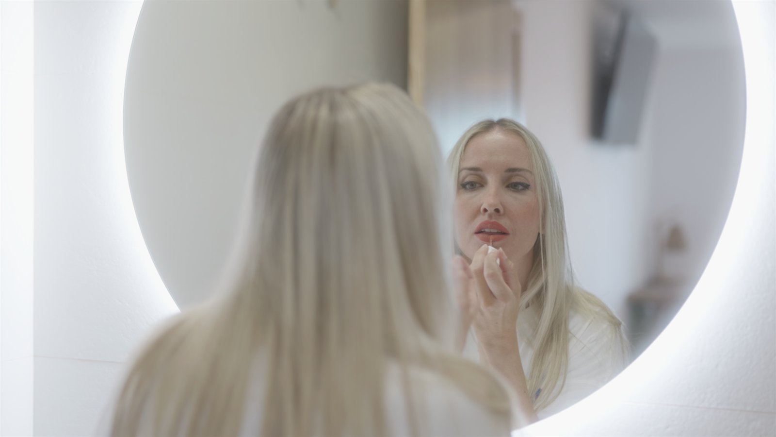 Mujer rubia aplicándose maquillaje frente a un espejo circular iluminado.