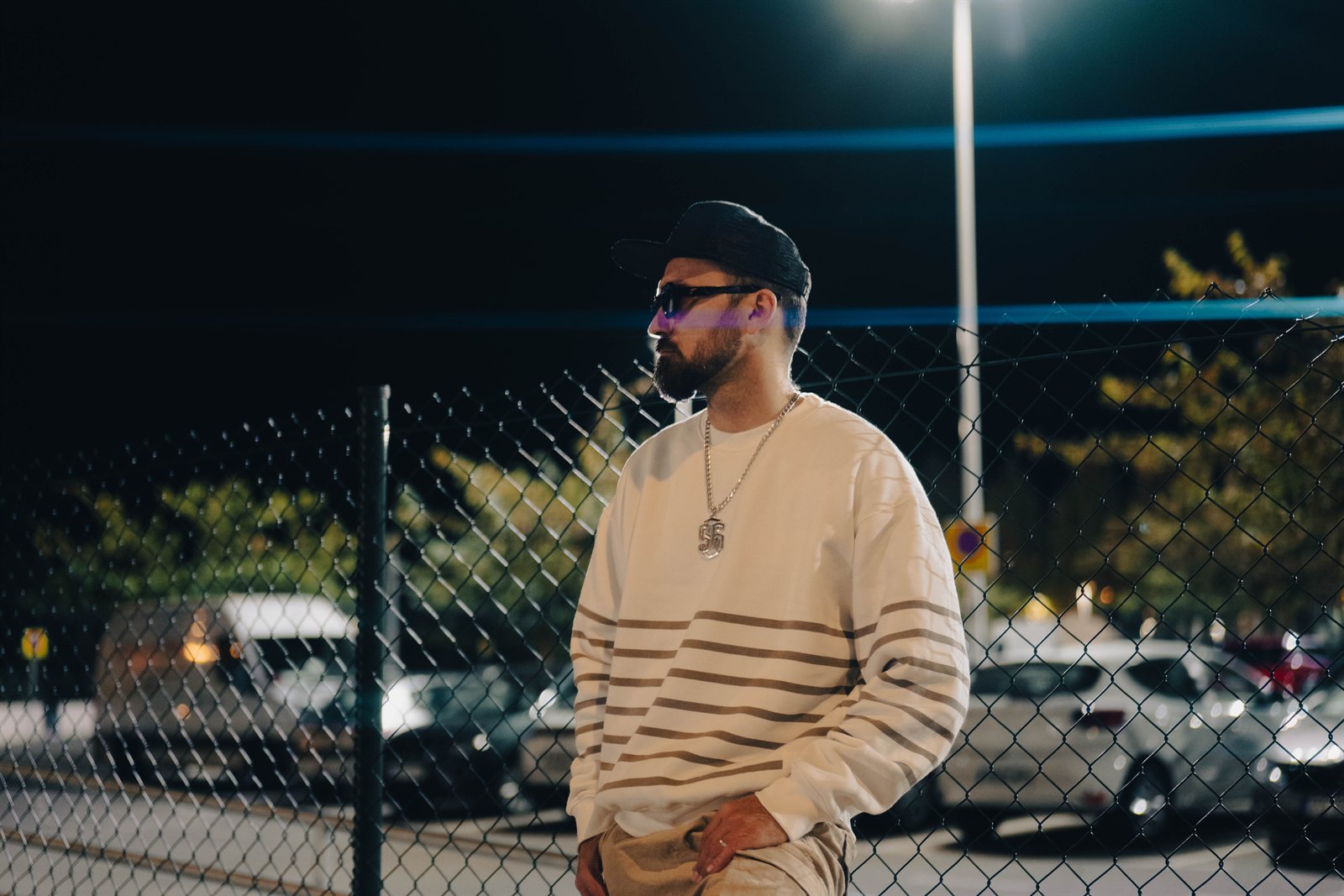 Retrato nocturno de un hombre con gorra y gafas junto a una valla metálica.