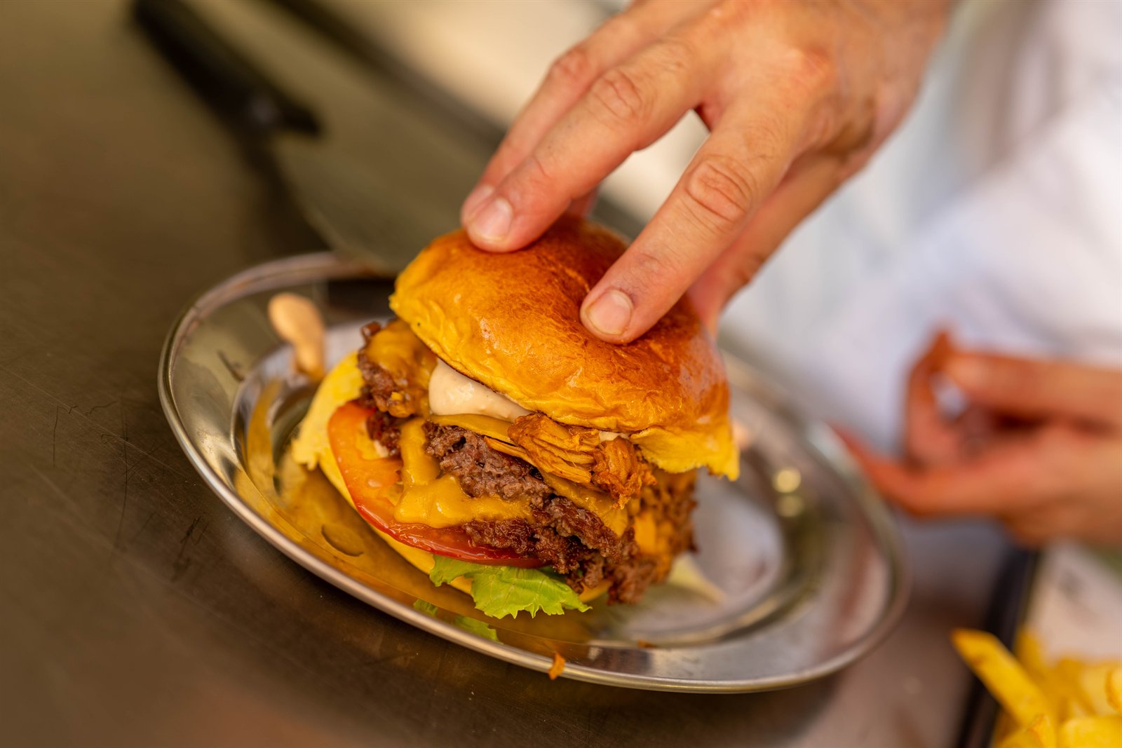 Mano terminando de montar una hamburguesa de varias capas sobre una bandeja metálica.